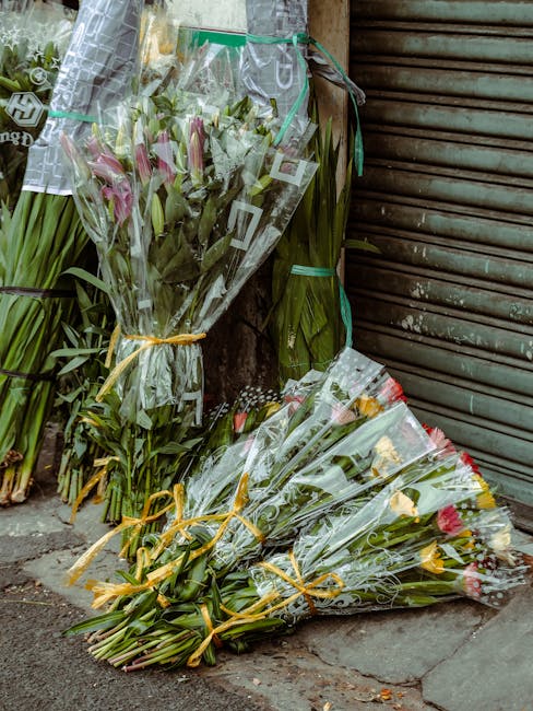 Several bouquets of fresh flowers, including pink lilies, yellow roses, and mixed greenery, wrapped in clear cellophane with gold and red ribbons, are arranged on a pavement outside a closed metal shu