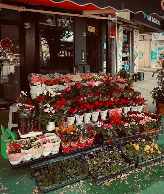 An outdoor display of fresh flowers in front of a florist shop, arranged on tiered black pallets and white tables. The arrangement includes vibrant red roses, pink tulips, white daisies, and purple pa