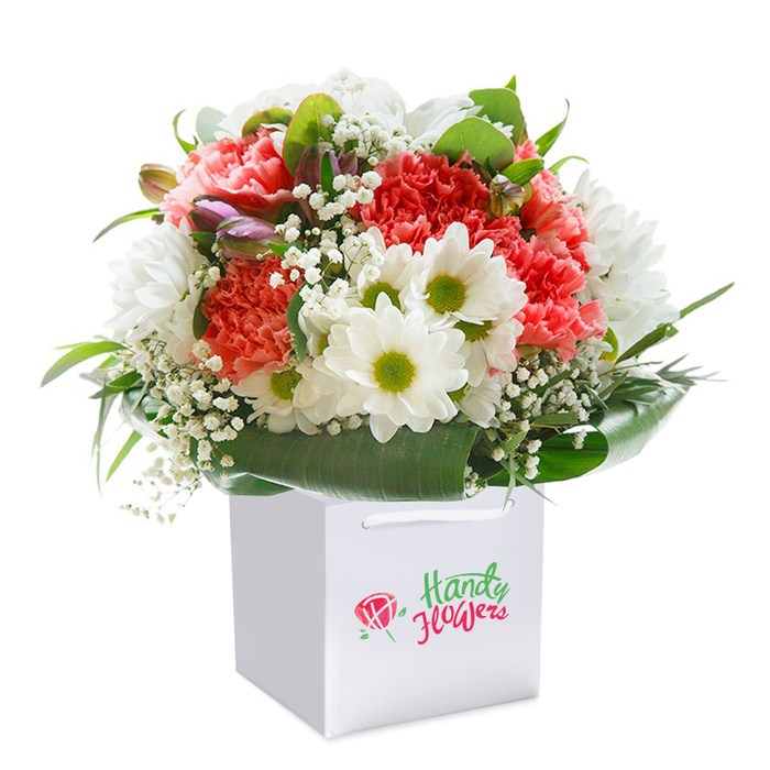 A beautifully constructed bouquet, brimming with colour and texture, sits proudly in a crisp white square gift bag, its entirety set against a clean, luminous background. Prominent coral and peach carnations, recognisable by their plush, frilled petals, mingle with pure white daisy-like chrysanthemums whose striking lime-green centers evoke the vitality of a Saturday stroll through Stroud Green's local parks. Airy clouds of white baby's breath are scattered strategically to add lightness throughout, while a few slender, purple-tinged buds-perhaps closed alstroemeria or similar-peek from the left edge, introducing depth and visual intrigue. Lush, deep green leaves fan outward in glossy arcs, forming a natural collar that grounds the composition, much like the mature street trees lining the quiet avenues of Stroud Green. The gift bag's square structure and strong white ribbon handles echo a modern sensibility, ideal for delivering heartfelt surprises to friends gathered at nearby cafés or family gatherings on Mount View Road. Every floral choice, every gentle transition between hue and texture, has been arranged with a local florist's discerning eye, ensuring a rounded, abundant form that radiates freshness and intention. This bouquet is more than an arrangement-it's a reflection of Stroud Green's sense of celebration and care, suited for birthdays, anniversaries, or moments when only the most thoughtfully prepared blooms will do.
